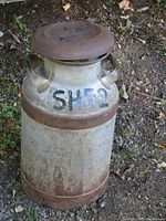 Front view of antique metal milk can standing outdoors on soil with rust patina and handle details visible.