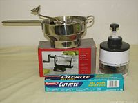 Full lot displayed showing all items: stainless steel food mill on a box of apple peeler with wax paper in front, and food chopper behind