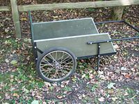 Left side profile view of the dock cart showing the large spoked wheel, green wooden panels, and metal handlebar resting on leaf-covered ground.