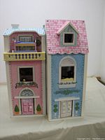 Front view of closed dollhouse showing two attached sections with pink and blue exteriors, decorative window boxes, doors and balcony.