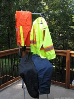 Overview of four men's work clothing items hanging on a metal rack outside: bright orange safety bib overalls, neon yellow safety coat, black insulated pants, and blue Stihl chain saw protective pants.