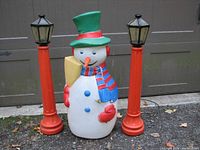 Front view of the plastic snowman figurine standing between two tall red lamp posts with black lantern tops, showing details and colors.