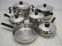 Overview of seven stainless steel pots and pans, all with lids and black handles, arranged on a white surface.