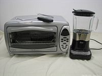 Front view of silver Europro rotisserie oven with glass door and Oster blender side by side on a white surface.