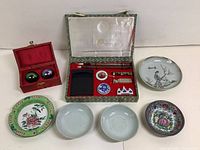 Photo showing all items: calligraphy set in green box, baoding balls in red box, and five small ceramic plates with various Asian designs.