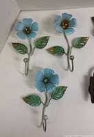 Three white metal floral wall hooks with gold accents and green leaves
