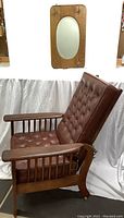 Side view of the Morris chair with brown tufted faux leather cushions and the oak mirror with brass hardware mounted on the wall behind it.