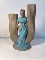 Front view showing the woman figure in blue dress holding a flower, flanked by two tall gray tube vases.