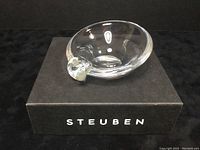 Steuben crystal ash receiver shown sitting on its storage box, clear and polished with a protruding molded detail on one edge.