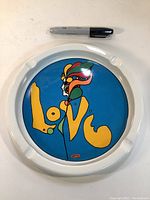 Top view of large white ceramic ash tray with colorful Peter Max 'Love' design and a Sharpie marker for scale