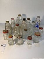 Front view of 16 assorted clear glass jars with metal lids, arranged in several rows showing variety in shapes and sizes, many bear-shaped and featuring coin slots in lids.