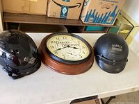 Wall clock centered with glossy black helmet on left and matte black helmet on right on table.