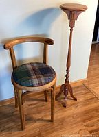 Wide view showing the wooden accent chair with curved back and plaid cushioned seat next to the tall wooden pedestal plant stand.
