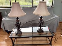Photo showing the two tier rectangular glass table with black metal frame and the pair of ornate wrought iron lamps with beige fabric shades sitting on top.