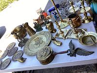 Full view of brass and brass-plated decorative items arranged on table, showing plates, candlesticks, bust cup, bookends, eagle sculpture, and small knickknacks.