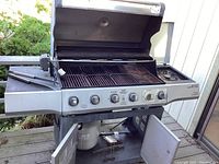 Front view of the stainless steel Brinkmann grill with hood open, showing five grilling grates and control panel with six knobs. Lower cabinet door open revealing propane tank.
