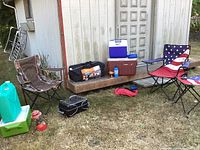 Camping setup on grass with two folding chairs, coolers, lantern, water container, portable cook stove, and duffel bag with tent