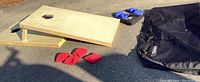 Photo showing two wooden corn hole boards with circular holes, four red bean bags, blue bean bags to the side, and a large black carrying bag on asphalt.