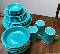 Photo showing stacked turquoise plates (dinner, sandwich, saucers) and some bowls with 3 mugs upside down showing Fieste USA marks.