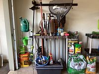 Lot of gardening supplies including Ryobi weedwhacker, hand tools on rack, watering cans, bags of fertilizer and plant food, and garden accessories arranged in garage.
