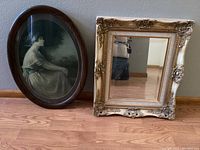 Both items side by side on wooden floor: gold ornate mirror and oval framed print.