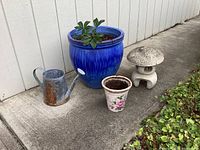 Set of four garden items: cobalt blue ceramic planter with plant, small ceramic rose pot, rustic metal watering can, stone Buddha statue