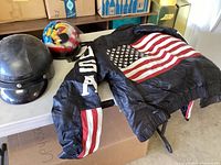 Leather jacket with two helmets displayed on a table. The jacket features an American flag on the back and USA lettering on the sleeve. The helmets include one with graphic designs and one plain black.