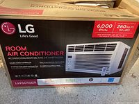 Original sealed box of the LG LW6015ER Room Air Conditioner showing the front label with key specifications including 6000 BTU/hr cooling capacity, suitable for 260 sq.ft. rooms, R410A refrigerant, and 1 year warranty.