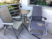 Reclining chaise lounge, small square smoked glass and metal patio table with 2 folding patio chairs on wooden deck.