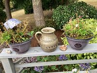 Plum plastic plant containers with flower plants, ceramic pitcher, mushroom, and decorative rocks on a gray wooden bench.