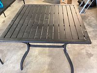 Square metal table with slatted top and curved legs on concrete floor.