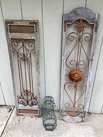 Blue metal wire bird cage and two distinct wrought iron decorative panels placed against a wooden wall, showing full view of all items.