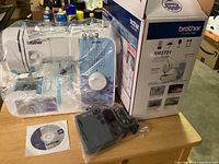 Brother XM2701 sewing machine set up on table with foot pedal and CD manual, still in plastic wrap, next to the original box showing machine model.