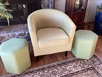 Front view of pistachio fabric barrel chair flanked by two pistachio hexagon stools on hardwood floor with area rug, fireplace, and furniture in background.