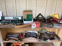 Overview of lot items including socket wrench set in green case, assorted hand tools, hammer, squares, and staples boxes on wooden shelves.