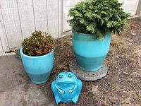 Wide shot of two teal ceramic pots with plants and ceramic frog figurine in driveway on grass and paved area.