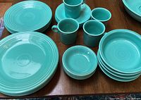Photo showing full set of turquoise Fiesta dinnerware including plates, mugs, saucers, and bowls arranged on wooden table.