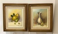 Two framed oil on canvas paintings by R. Monti placed side by side on a white wall. One features pear and grapes, the other a silver vase and grapes.