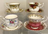 Four Royal Albert bone china teacups with matching saucers, displayed on clear stands showing diverse floral patterns and gold trim.