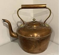 Photo of copper teapot showing side angle with handle, lid, and spout on table against wall.