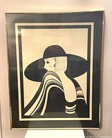 Framed black and white art deco print featuring a woman in a large hat and striped shawl, showing the overall item and condition.
