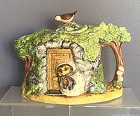 Front view of the teapot showing the hedgehog character at the door of a cottage surrounded by greenery.