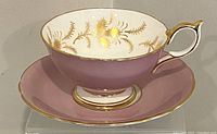 Front side view of the Aynsley tea cup sitting on the matching saucer showing the mauve exterior and gold floral pattern inside.