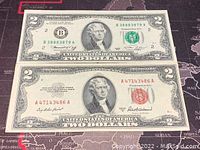 Photo showing both 1953 and 1976 US two dollar banknotes placed side by side on fabric with world map print.