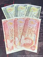 7 Canadian banknotes displayed, three 1973 1-dollar notes and three 1974 2-dollar notes spread out on black surface.