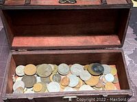 Open wooden chest containing a mixed assortment of coins of various sizes and colors including silver, bronze, and copper tones.