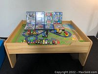 Full view of wooden Thomas the Train play table with track print surface, toy trains, and DVDs placed on top.