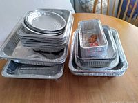 Stacked aluminum disposable steam table pans and foil trays on wooden table, various sizes.