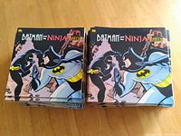 Two piles of 'Batman and the Ninja' comic books resting on a wooden surface. Each comic features the same cover showing Batman facing a ninja.