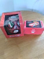 Photo showing both Coca-Cola items in their original boxes. The soccer ball box has a clear plastic window and is damaged; the bowl-a-rama bank box is red and less visible.